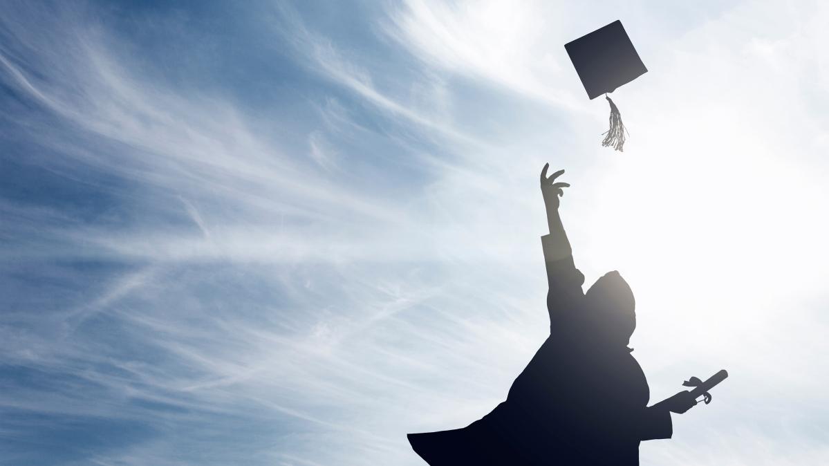 Silhouette of a person in a graduation gown holding a diploma and throwing their tassled hat in the air against a blue, cloud painted sky.